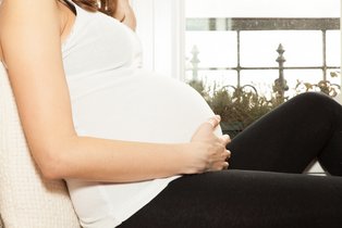 Eine schwangere Frau mit weißem Hemd und schwarzer Hose sitzt auf einem Sofa und hält ihren Bauch von unten fest Kopf und die kompletten Beine sind zu sehen